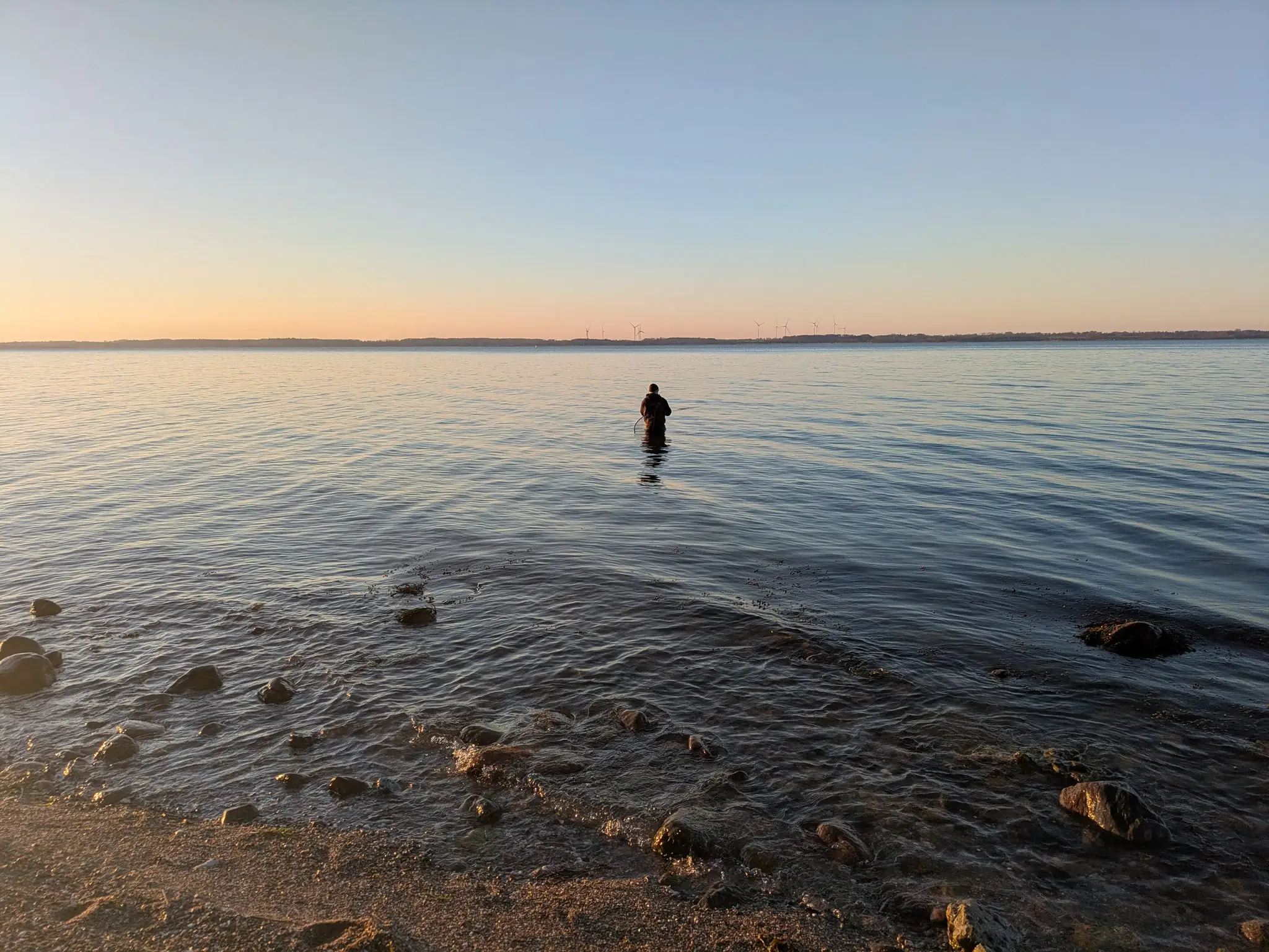 Angler watet bei Sonnenuntergang ins ruhige Meer, Windräder am Horizont, Ostseeküste