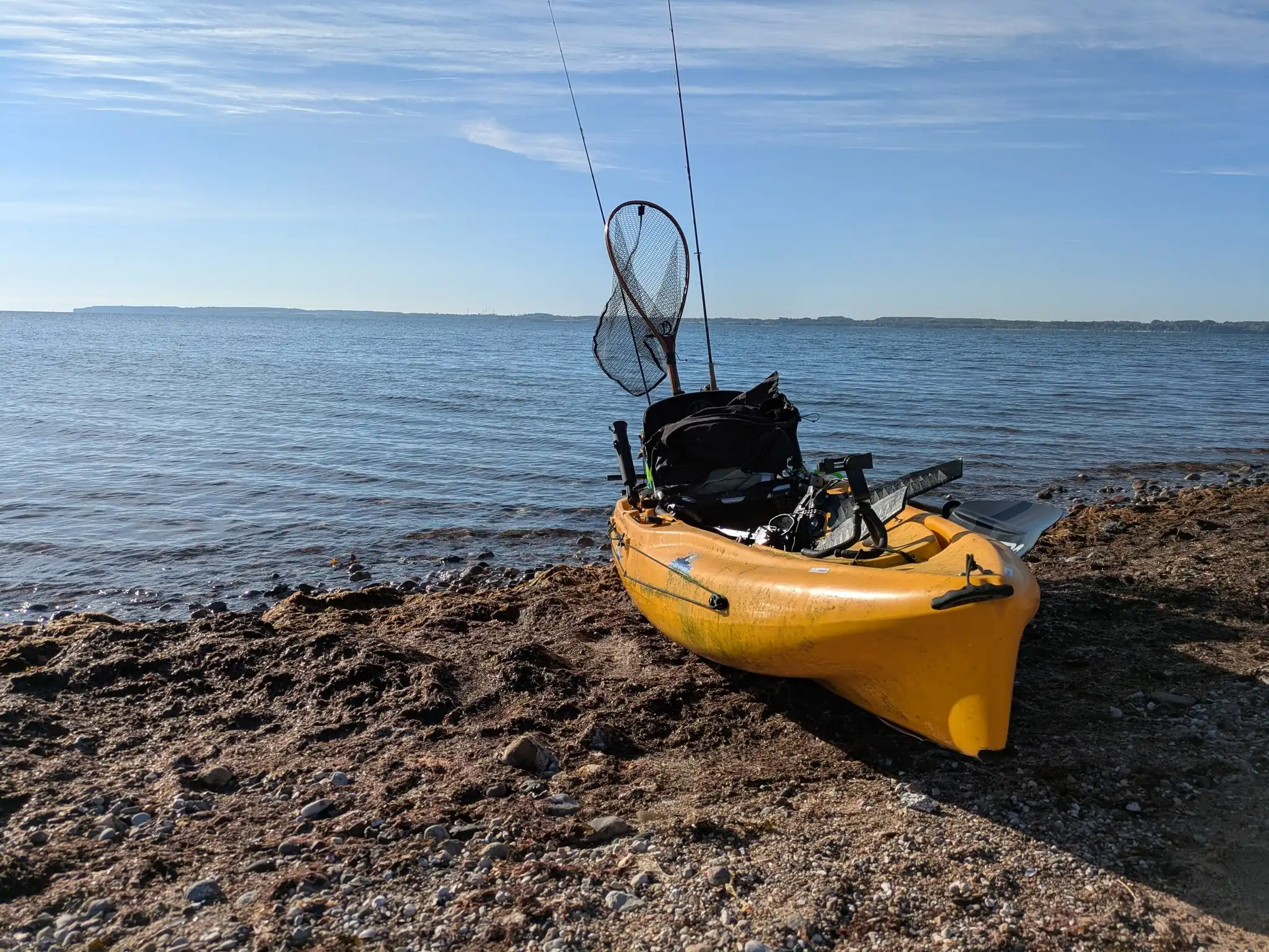 Gelbes Angelkajak mit Ruten und Kescher am Kiesstrand eines ruhigen Meeresufers