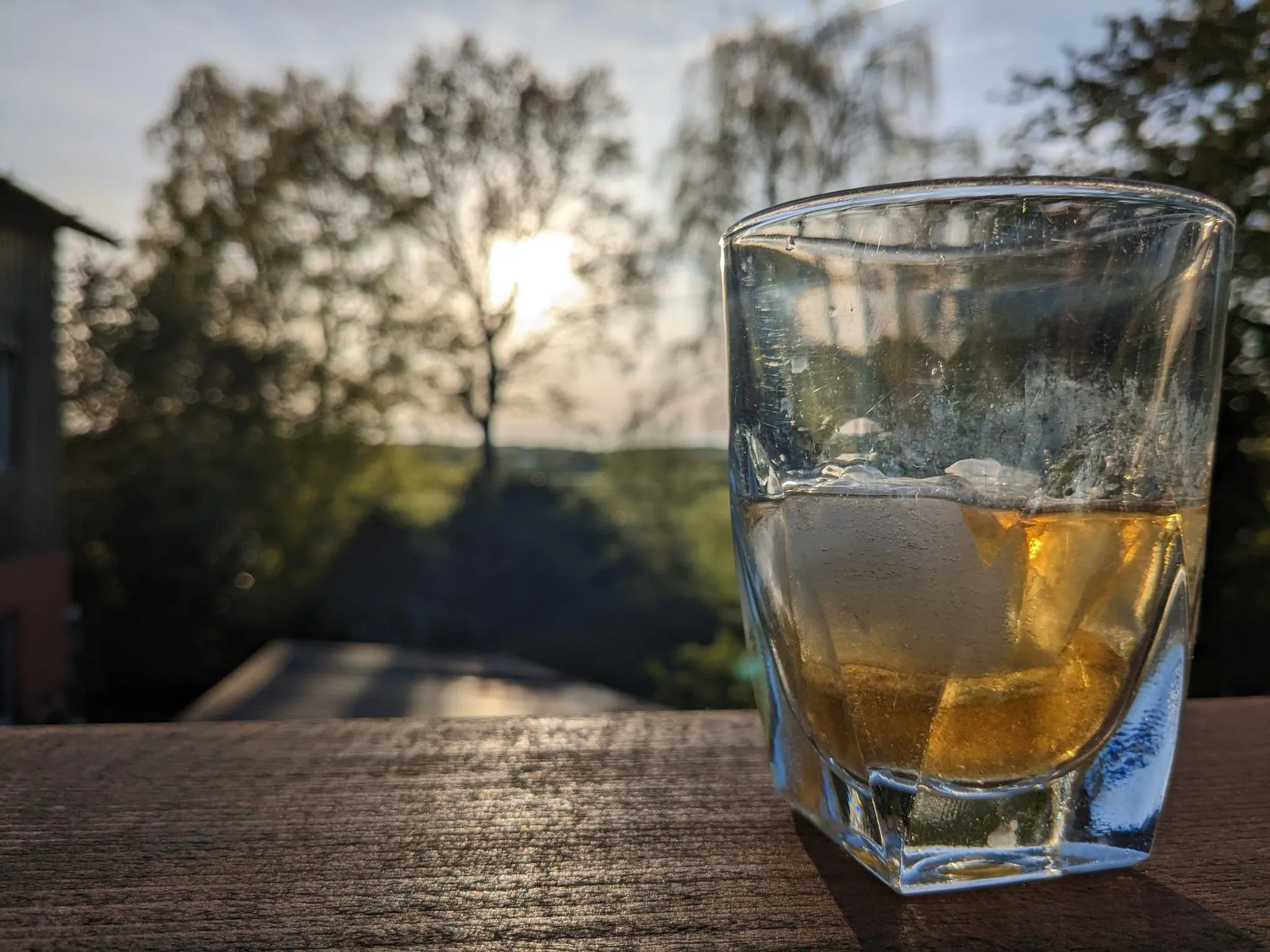 Whisky-Glas mit Eiswürfeln auf Holzgeländer bei Sonnenuntergang mit grüner Landschaft im Hintergrund