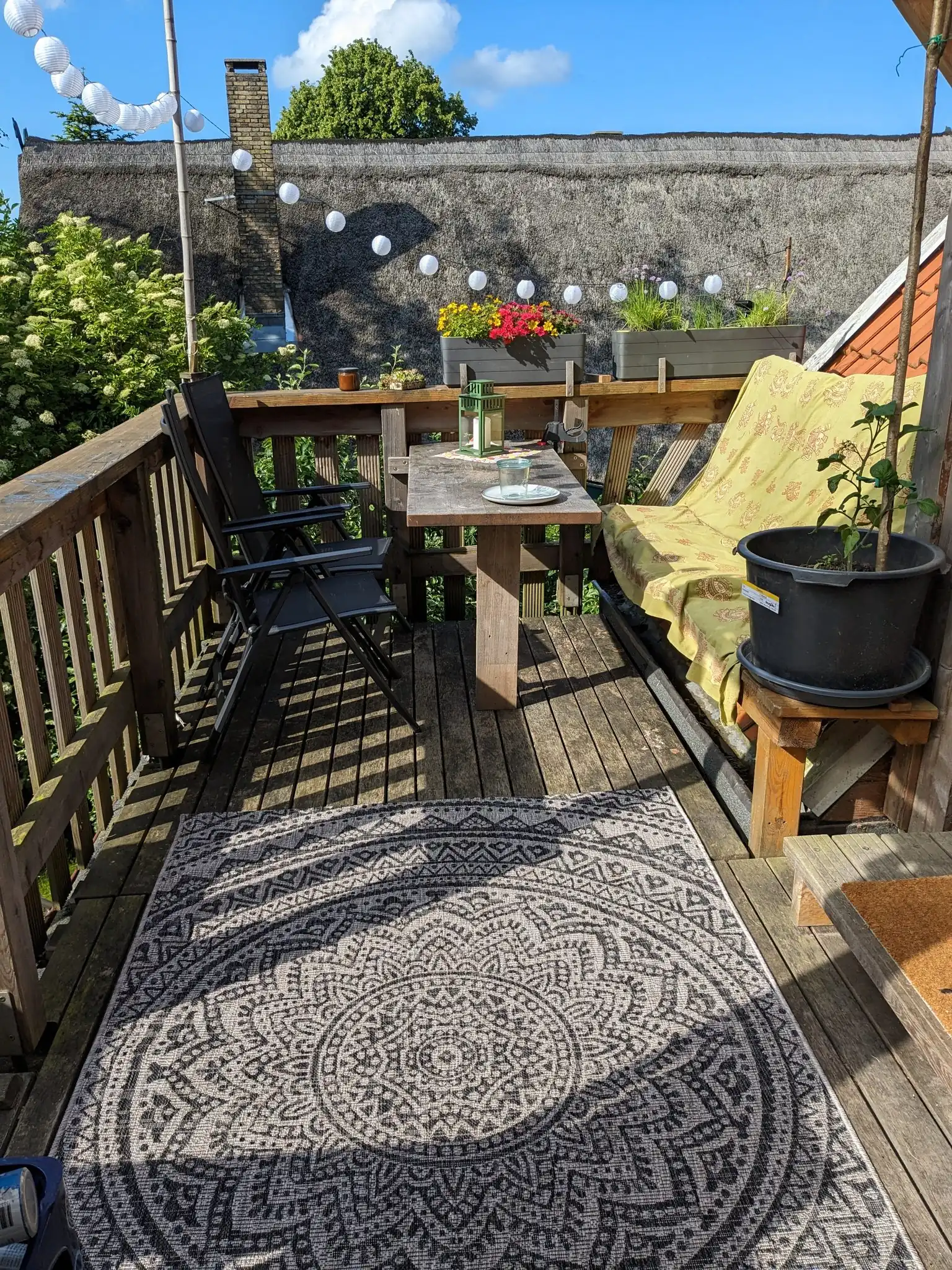 Sonnige Holzterrasse mit Esstisch, schwarzen Stühlen, Mandala-Teppich und Reetdach im Hintergrund