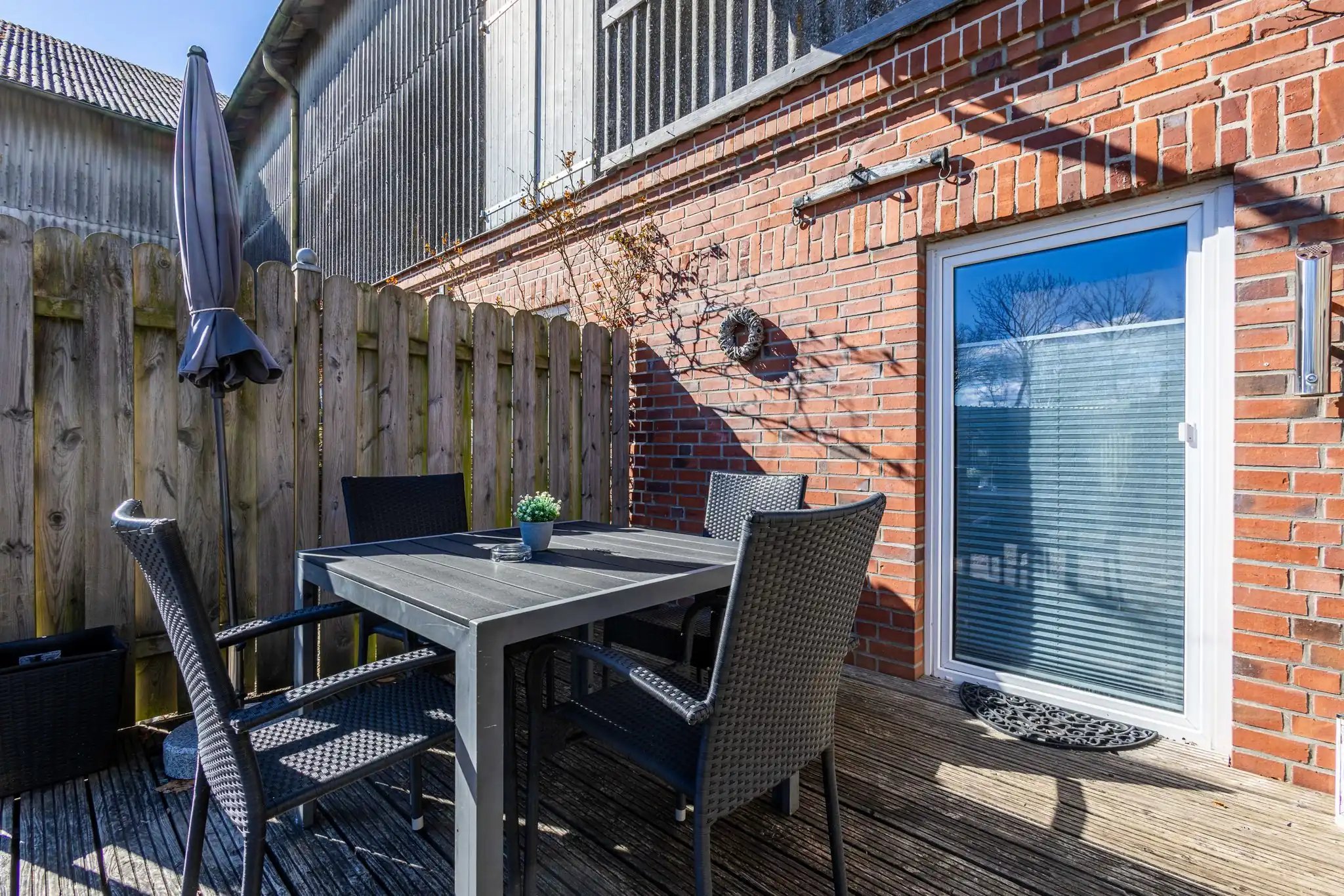 Sonnige Holzterrasse mit grauem Gartentisch, Rattanstühlen und Sonnenschirm an Backsteinhaus