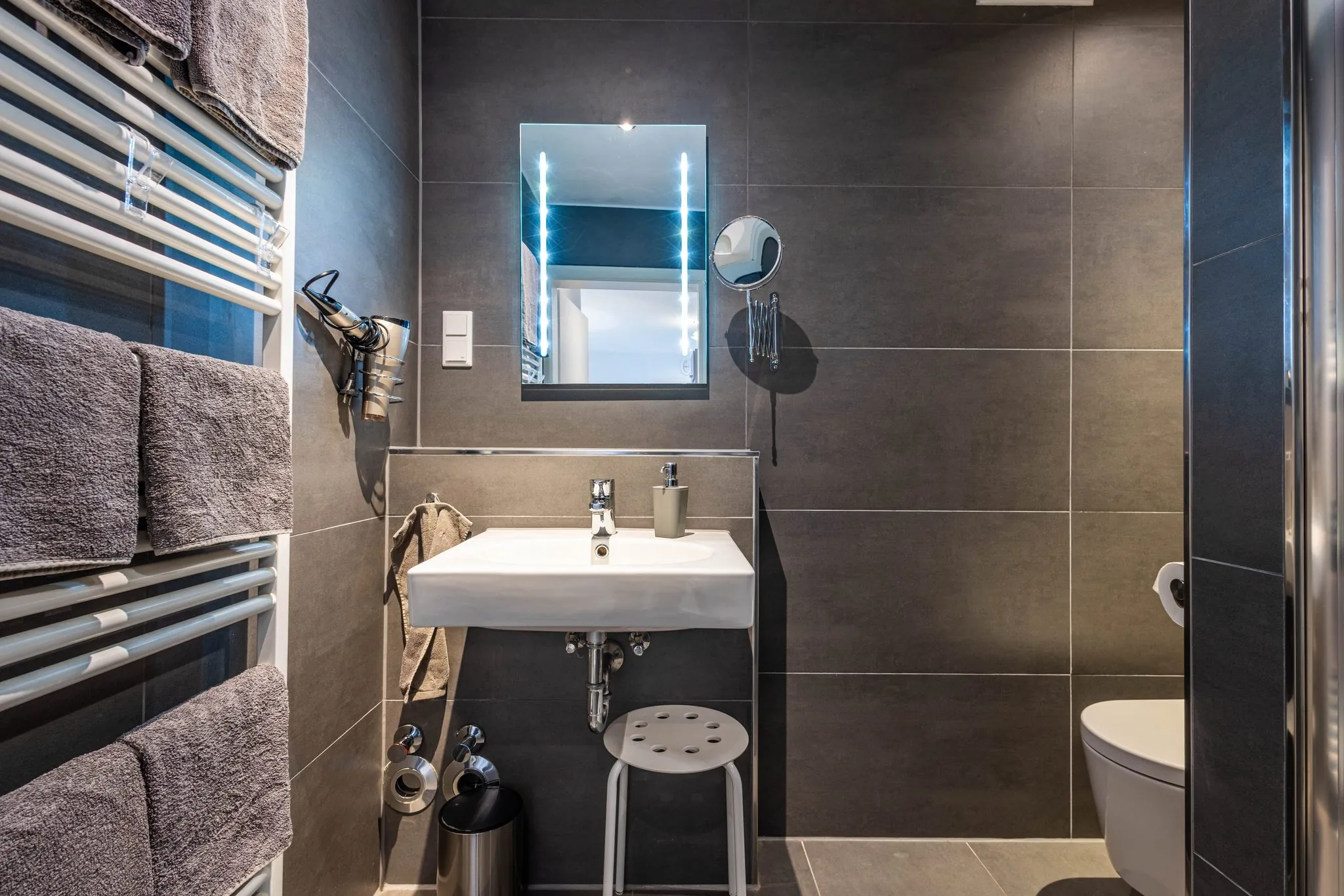 Modernes Badezimmer mit beleuchtetem Spiegel, Waschbecken, Handtuchheizung und Dusche in dunklen Fliesen