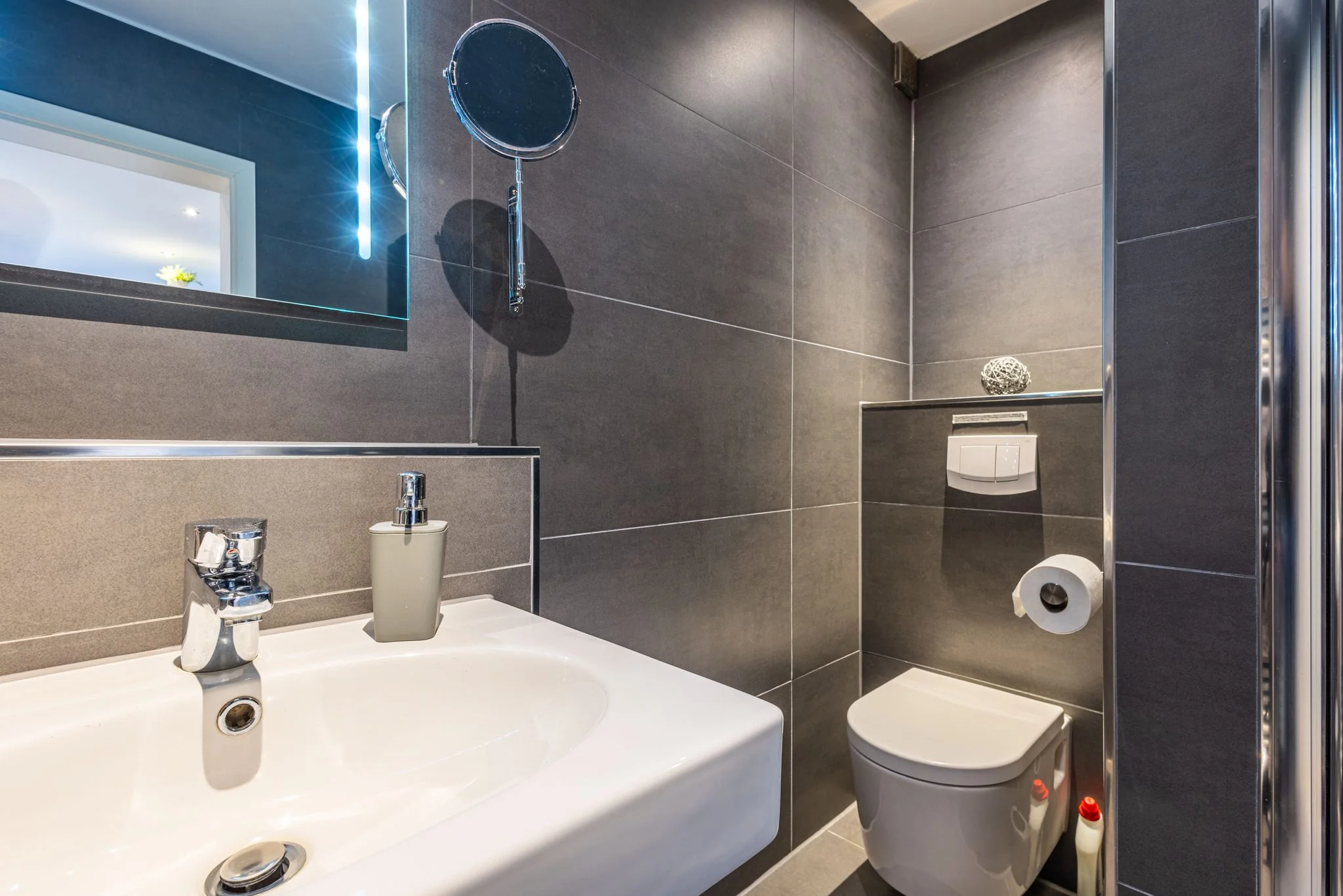 Modernes Badezimmer mit grauer Wandfliesen, Waschtisch, Wandtoilette und Kosmetikspiegel