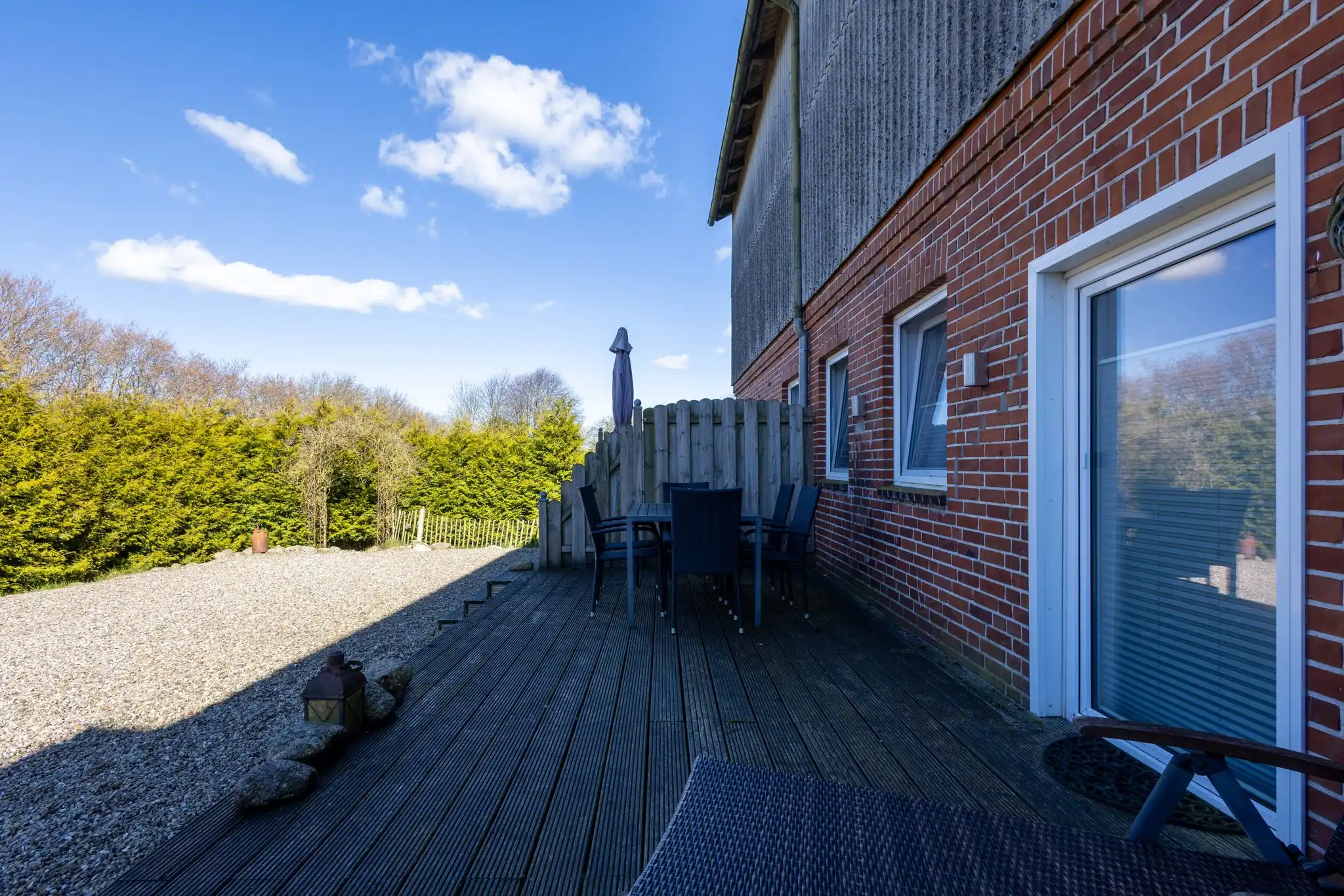 Holzterrasse mit Gartenmöbeln und Sonnenschirm an einem roten Backsteinhaus mit Kiesweg und Hecke