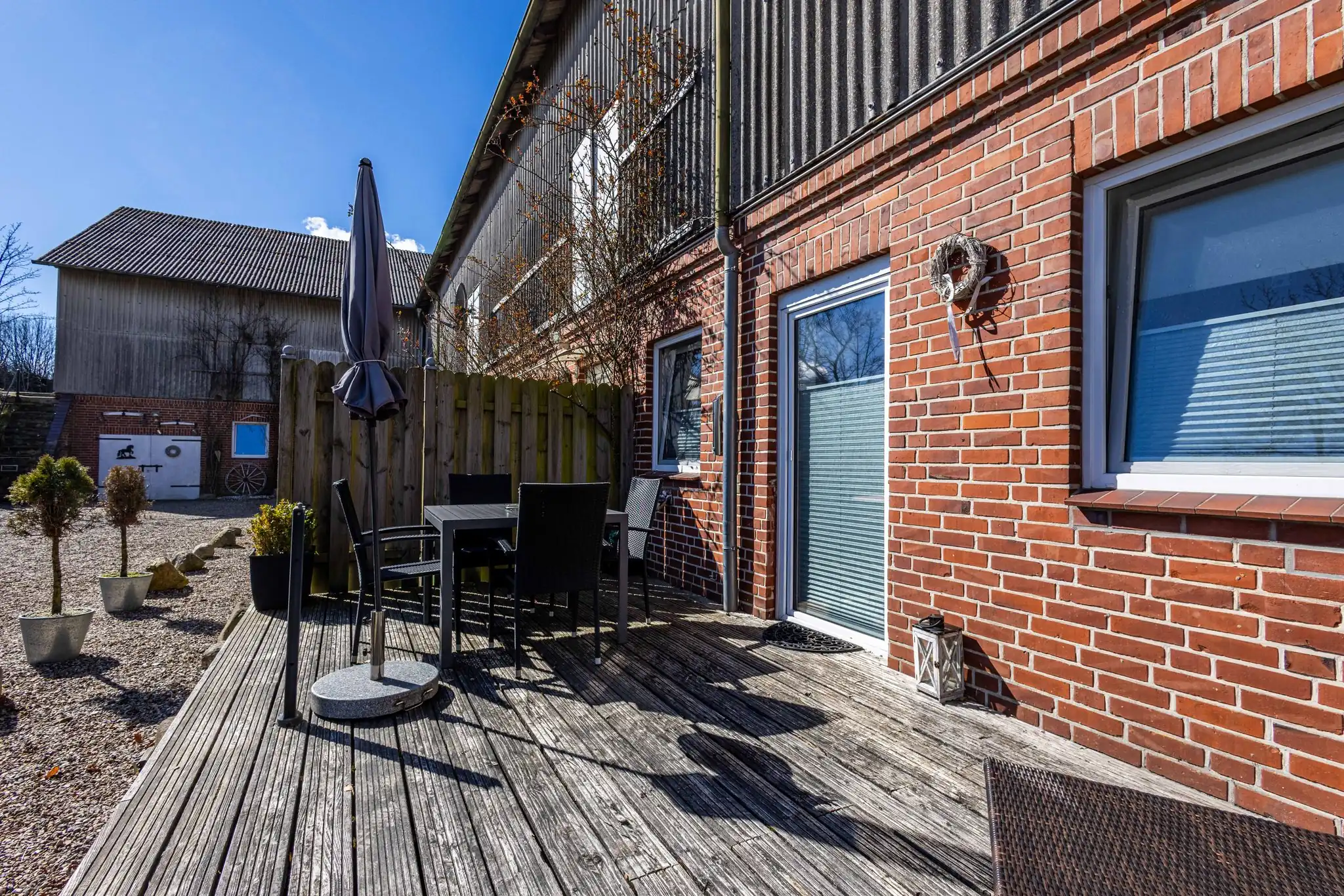 Holzterrasse mit Gartenmöbeln und Sonnenschirm an rotem Backsteinhaus auf Bauernhof
