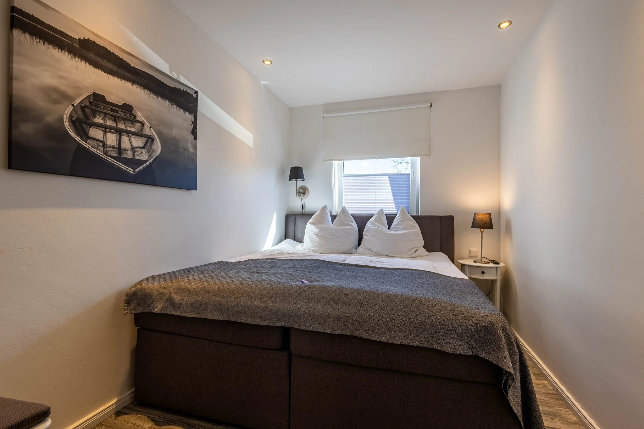 Gemütliches Schlafzimmer mit Doppelbett, grauer Tagesdecke und Wandbild eines Ruderboots