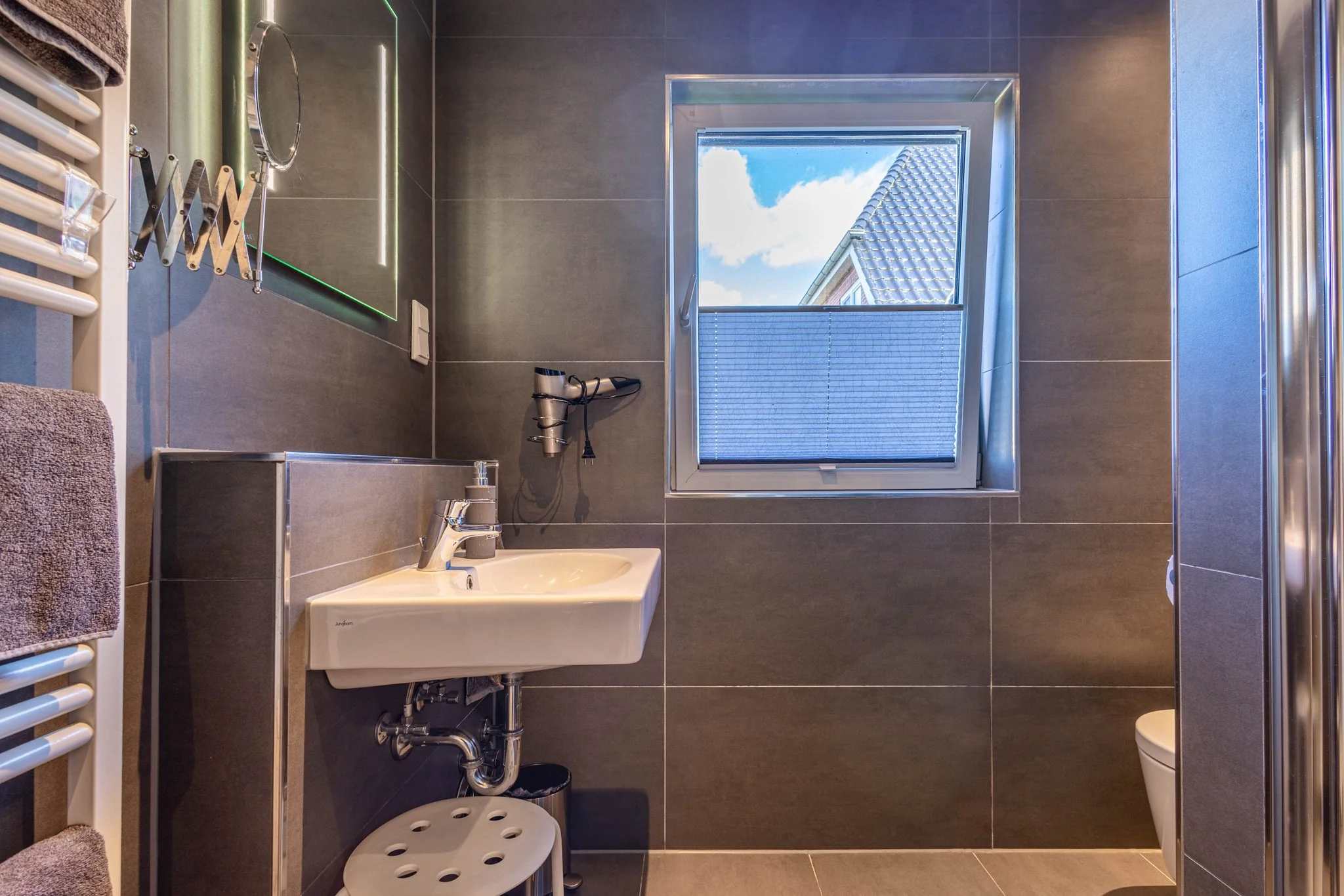 Modernes Badezimmer mit Waschbecken, Handtuchheizkörper, Spiegel und Fenster in grauer Fliesenoptik