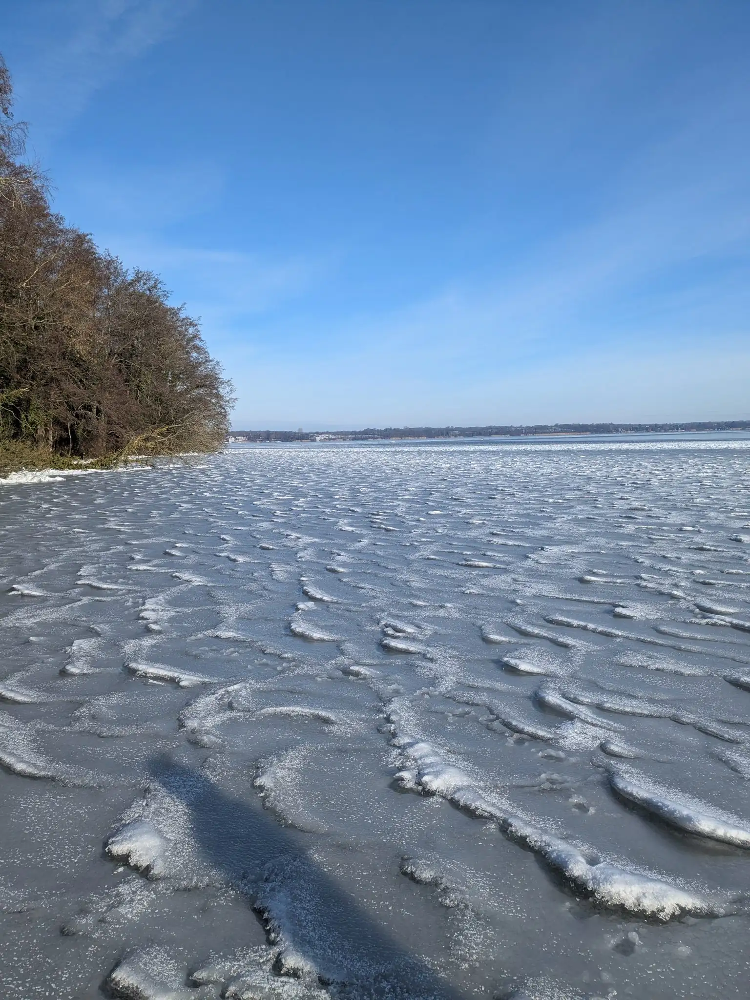 Zugefrorener See mit Eisschollen und schneebedecktem Ufer unter blauem Winterhimmel