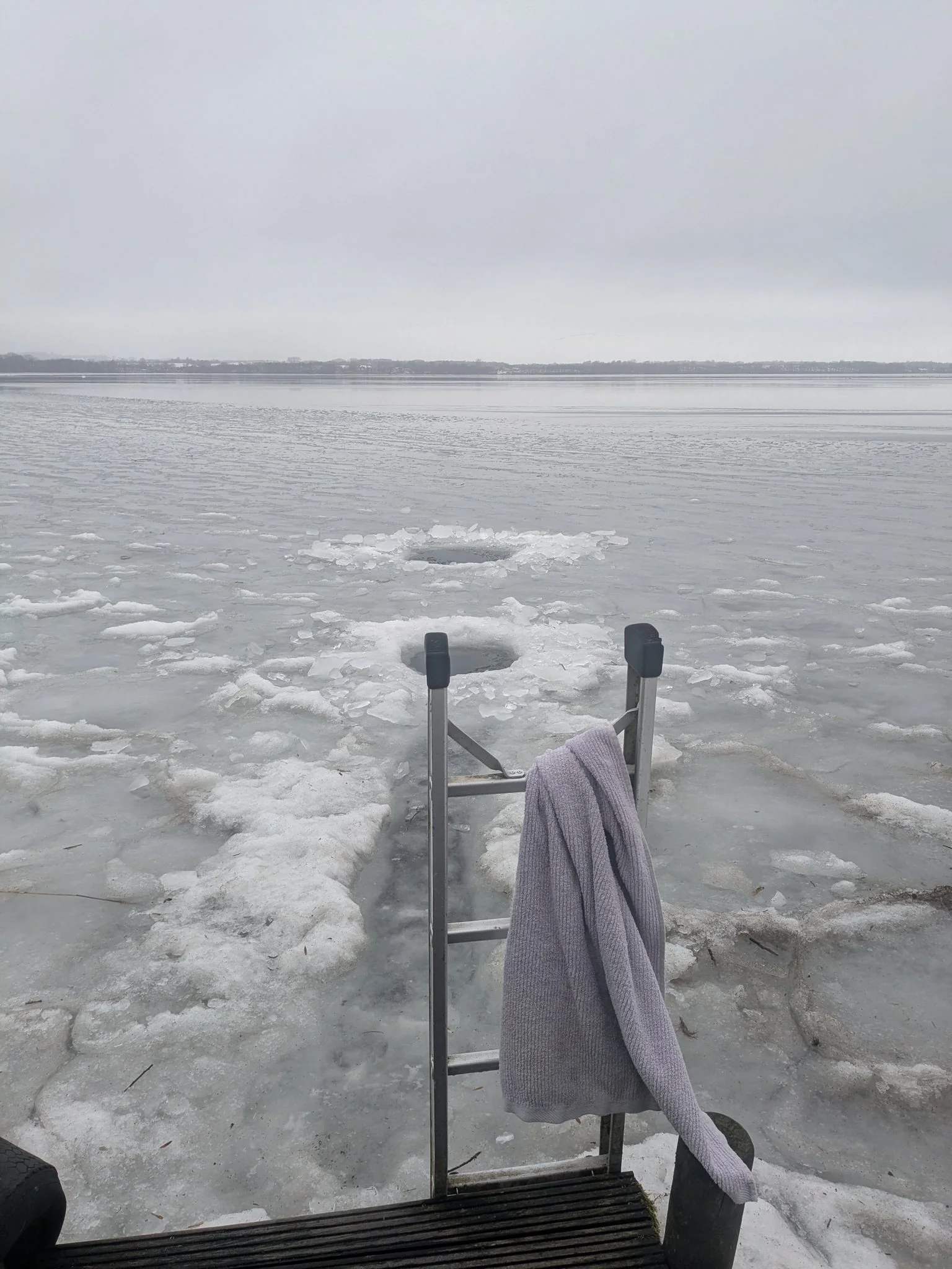 Eisbaden am zugefrorenen See mit Leiter, Eisloch und Handtuch im Winter